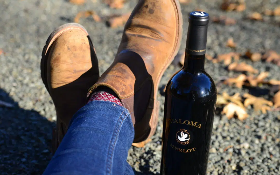 A bottle of 2018 Paloma Merlot beside a pair of crossed legs with boots on the feet.
